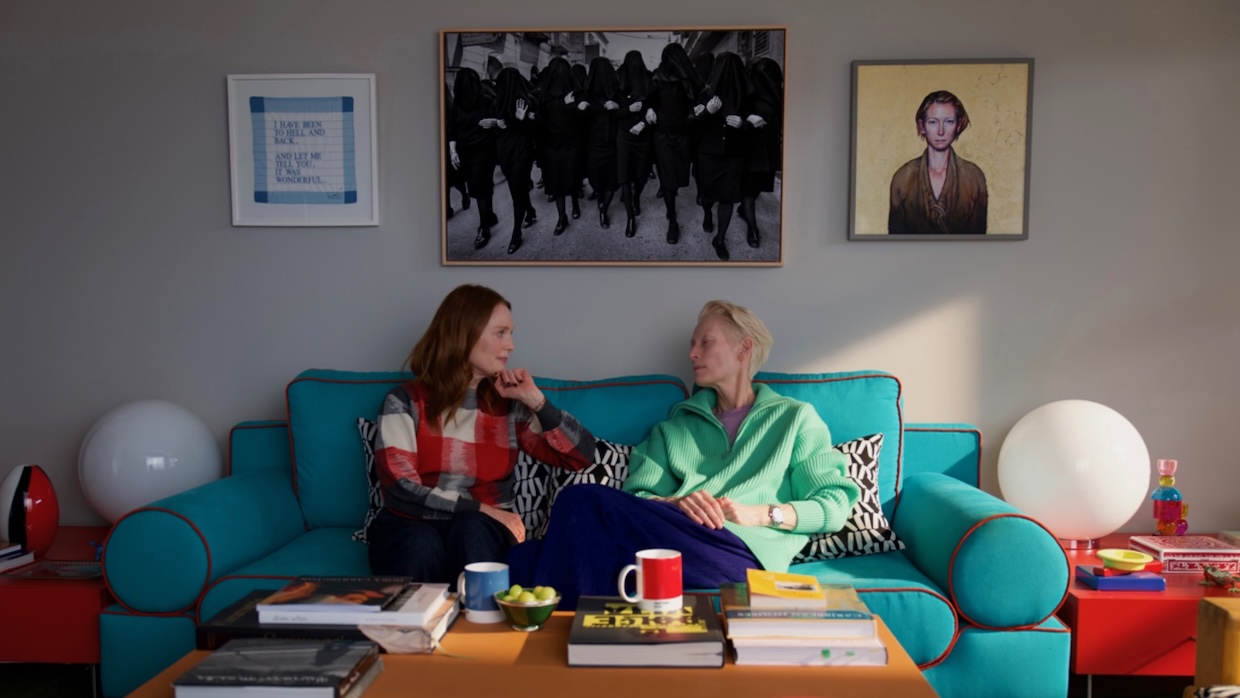 Two women sit on a bright blue couch in a sunny living room.